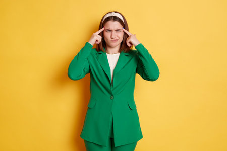 Sad woman wearing green jacket standing isolated over yellow background thinking trying to remember something frowning face keeps fingers in head.の写真素材