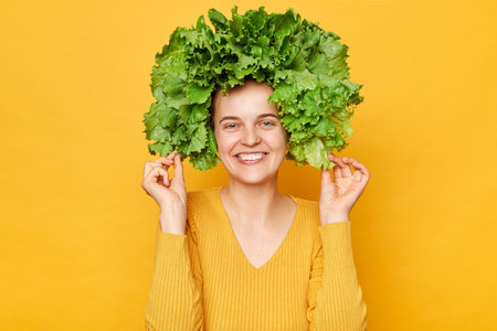 Positive woman wearing casual shirt and lettuce wreath standing isolated over yellow background looking at camera expressing positive emotions smiling toothily.の写真素材