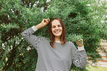 Portrait of happy smiling cheerful woman wearing striped shirt standing with raised arms and satisfied facial expression near green bush in park having great time outdoor.の写真素材