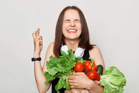 Hopeful adult woman with brown hair standing isolated over white background holding vegetables crossing her finger closing eyes making wish praying for good luck.の写真素材