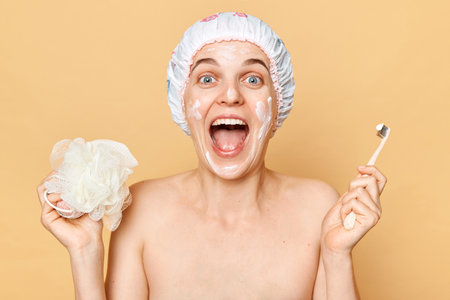 Morning hygiene and beauty procedures. Amazed woman holding toothbrush and washcloth wearing shower cap waking up in good mood has joyful expression.の写真素材