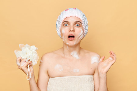 Shocked astonished surprised female dressed bath cap looking at camera with open mouth holding sponge with foam taking shower in thew morning expressing fear.の写真素材