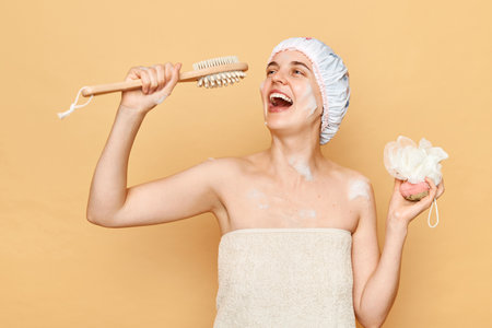 Playful positive woman taking shower in bathroom holding washcloth and body brush for scrub and massage like microphone singing happily having fun while doing skin care procedures.の写真素材