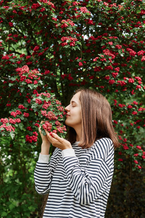 Joyful satisfied caucasian woman dressed striped shirt posing blooming tree in park in early spring nature enjoying pleasant aromat smelling flowers.の写真素材