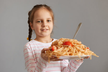 Cute smiling little kid girl with pigtails eating spaghetti holding big plate with pasta looking at camera waiting for dinner being extremely hungry.の写真素材