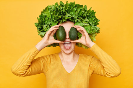 Nutritious food choices. Amazed excited woman with lettuce hair style with green vegetables on her head isolated over yellow background covering eyes with avocado screaming wow.の写真素材
