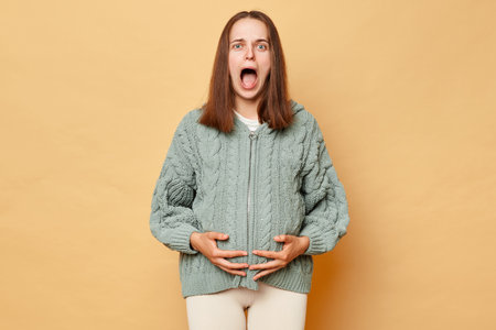 Shocked scared pregnant woman wearing knitted warm sweater standing isolated over beige background touching her belly screaming with fear feeling contractions.の写真素材