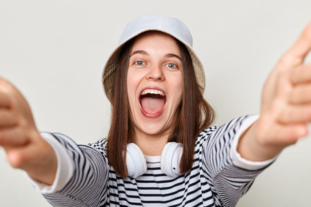Joyful extremely happy woman wearing striped shirt and panama standing with headphones over neck isolated over gray background making point of view photo screaming with happiness.の写真素材