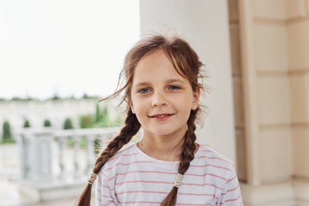 Outdoor shot of sweet charming cute little girl smiling looking at camera wearing striped shirt and having pigtails spending time alone expressing positive emotions.の写真素材