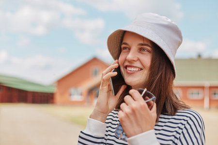 Portrait of happy attractive young caucasian young woman talking with cellphone while walking outside looking away with smile and having pleasant conversation.の写真素材