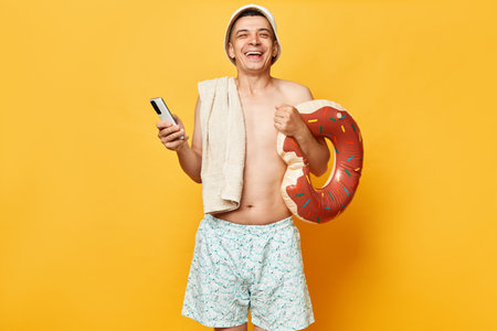 Laughing adult man wearing shorts swimsuit and panama holding donut rubber ring and bag isolated on yellow background using phone while resting at resort.の写真素材