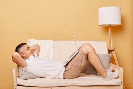 Smiling handsome man lying on sofa at home watching movie or film on laptop in the living room having satisfied expression being calm and relaxed keeps hands behind head.の写真素材