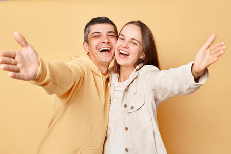 Friendly smiling woman and man wearing casual style clothing standing isolated over beige background spreading hands inviting showing welcome gesture laughing with happiness.の写真素材
