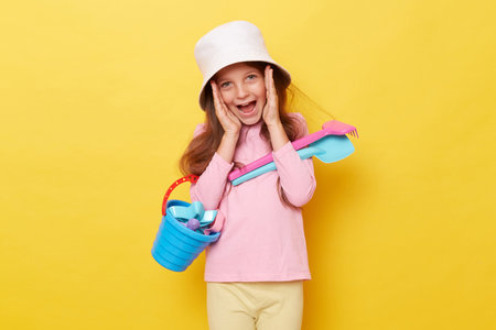 Excited little girl wearing casual clothing and panama holding sandbox toys and bucket isolated over yellow background screaming loud inviting friends to play together.の写真素材