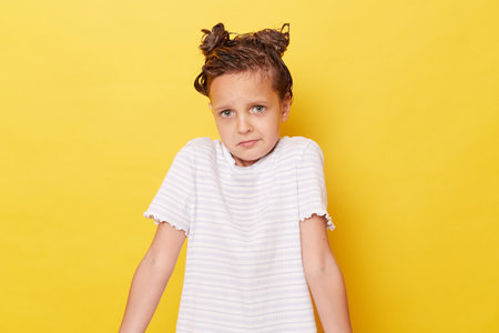 Uncertain little girl with wet hair wearing casual white T-shirt standing isolated over yellow background shrugging shoulders looking at camera with puzzled faceの写真素材