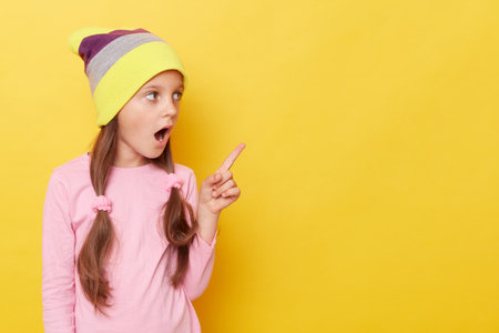 Shocked little girl with ponytails wearing beanie hat standing isolated over yellow background pointing away at advertisemet area being surprised.の写真素材