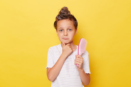 Puzzled child girl with shampoo on wet hair wearing white T-shirt standing isolated over yellow background holding comb looking at camera with confused expression suffering hair loss.の写真素材