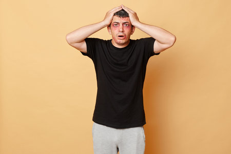 Shocked despair man in black T-shirt with bruises and abrasions on his face isolated over beige...