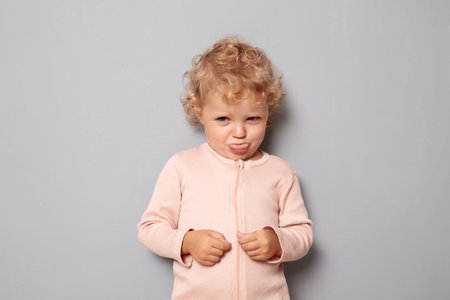 Funny xsad offended baby girl with blonde wavy hair wearing rose shirt isolated over gray background...