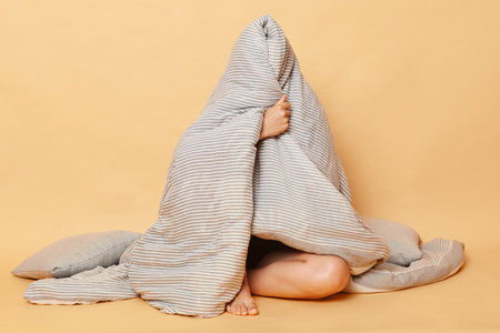 Unknown unrecognizable woman sitting on floor wrapped in plaid wearing pajama, sleep eye mask isolated over beige background hiding herself in blanket.の写真素材