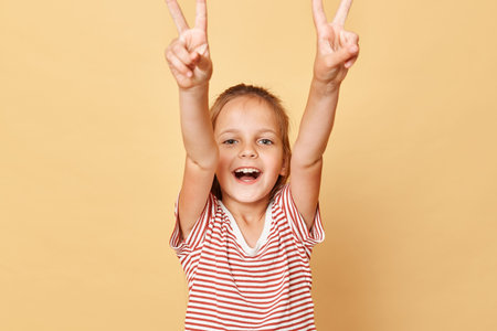 Excited smiling dark haired little girl wearing striped T-shirt isolated over beige background showing victory sign screaming with happiness rejoicing.の写真素材