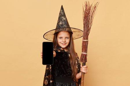 Delighted charming little girl with long hair wearing black witch costume and carnival hat cone holding broom isolated over beige background showing smartphone with empty screen for advertisement.の写真素材