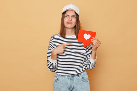 Unhappy woman wearing striped shirt and baseball cap holding pointing at like blogger icon standing...