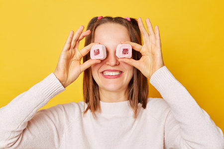 Satisfied happy unknown young girl with ponytails in jumper covering her eyes with marshmallow...