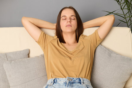 Calm young woman feeling fatigue sitting on comfortable sofa keeps hands behind head resting at home in her living room keeping eyes closed relaxing after hard day.の写真素材