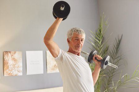 Strong confident gray-haired mature man wearing casual white T-shirt sitting on sofa at living room...