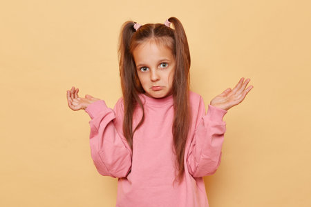 Puzzled uncertain little girl with ponytails wearing pink sweatshirt posing isolated over beige...