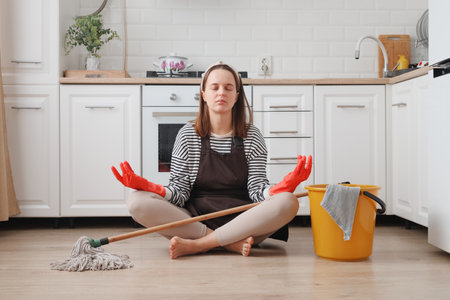 Tired woman wearing apron and rubber gloves housewife cleaning floor with steam mop in kitchen at home trying to relax and rest using yoga practice calming downの写真素材
