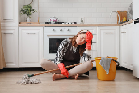 Exhausted woman wearing apron and rubber gloves housewife cleaning floor with steam mop in kitchen at home working long hours having headache sitting on floor with sad faceの写真素材