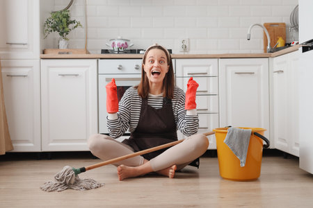 Overjoyed happy cheerful woman wearing apron and rubber gloves housewife cleaning floor with steam mop in kitchen at home clenched fist rejoicing to finish her household choresの写真素材