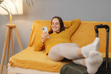 Beautiful cheerful Caucasian woman wearing orange sweater sitting with baggage on couch looking at mobile phone screen with smile browsing internet before leaving hotelの写真素材