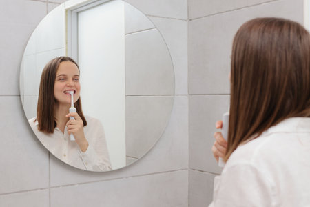 Clean and healthy mouth. Perfect oral hygiene. Dental protection methods. Morning oral care routine. Caucasian young adult woman brushing teeth in bathroom in front of mirrorの写真素材