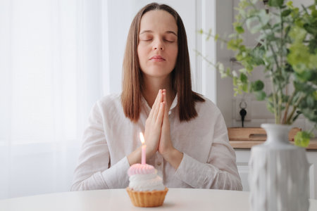 Calm attractive Caucasian woman in white shirt sitting at table with birthday cake and candle keeps...
