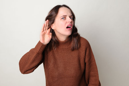 Caucasian woman wearing brown jumper standing isolated over gray background holding hand near ear...