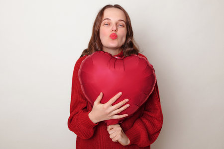 Romantic flirting brown haired adult woman wearing red sweater holding heart shape balloon sending air kissing with pout lips posing isolated over gray backgroundの写真素材