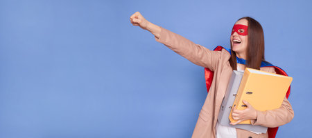 Confident strong successful brown haired woman wearing superhero costume holding paper folders posing isolated over blue background pretending mask flying with raised fist copy space for advertisementの写真素材