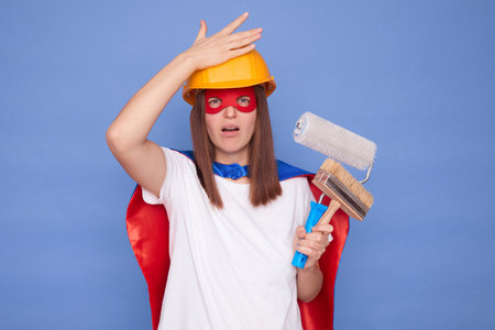 Regretful displeased woman painter wearing superhero costume and protective helmet holding painting roller and brush isolated on blue background showing facepalm gesture making mistake in paint mixingの写真素材