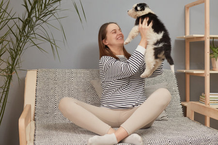 Satisfied happy smiling Caucasian woman wearing striped shirt sitting on the sofa raising her black...