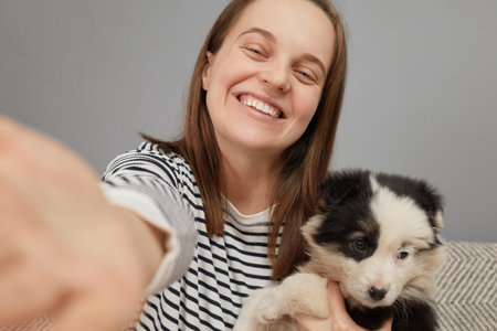 Smiling cheerful overjoyed woman in striped shirt sitting on sofa hugging her black and white puppy making point of view photo captures moment with new petの写真素材