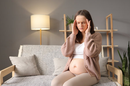Young pregnant woman suffering from headache while sitting on bed at home.の写真素材