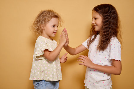 Wavy haired cheerful toddler clapping hands with her playful sister during active game of pat-a-cake both smiling carefree while cuddling together isolated on beige backgroundの写真素材