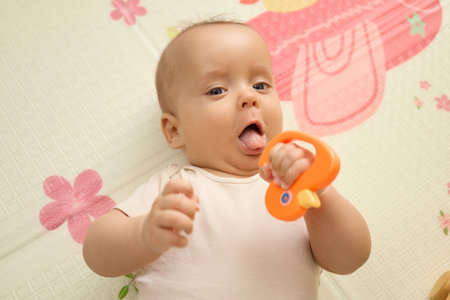 Top view of cute little Caucasian newborn baby boy wearing white bodysuit having teeth growing issues teething pain while holding bite toy lying on floor in living roomの写真素材