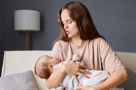 Brown haired woman loving mother holding her newborn baby in hands while sitting on couch gently...