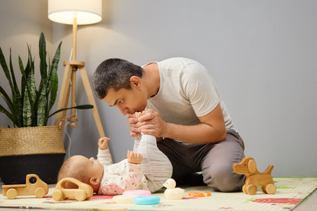 Loving cheerful father spending time with his newborn daughter or son on floor sharing moments of joy and happiness inside their home kissing tiny child's legsの写真素材
