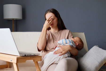 Exhausted woman rubbing her eyes while sitting on sofa with her baby balancing work deadlines and motherhood at her home workspace fatigued freelancer managing business tasks and parentingの写真素材