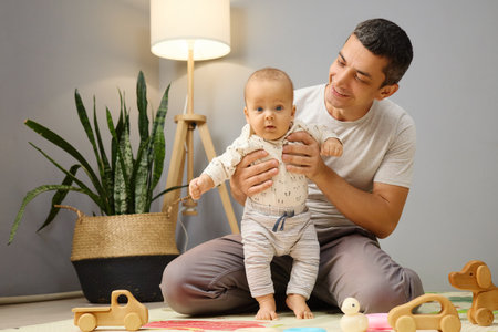 Positive attractive man playing with infant man sitting on floor indoors spending time together inside room father watching newborn developing holding child in handsの写真素材